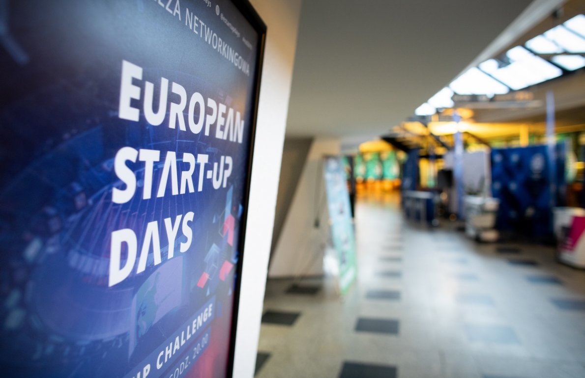 Niebieska tablica informacyjna European Start-up Days. W tle korytarz.